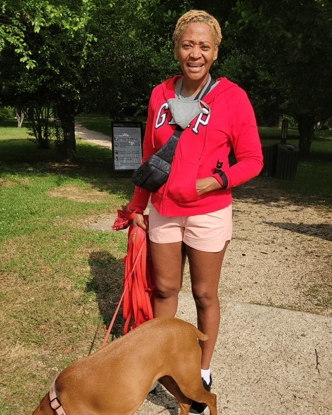 Photo of Junel Jeffrey in a red sweatshirt and shorts, smiling for the camera with her brown dog at the entrance to Kenilworth Aquatic Gardens.